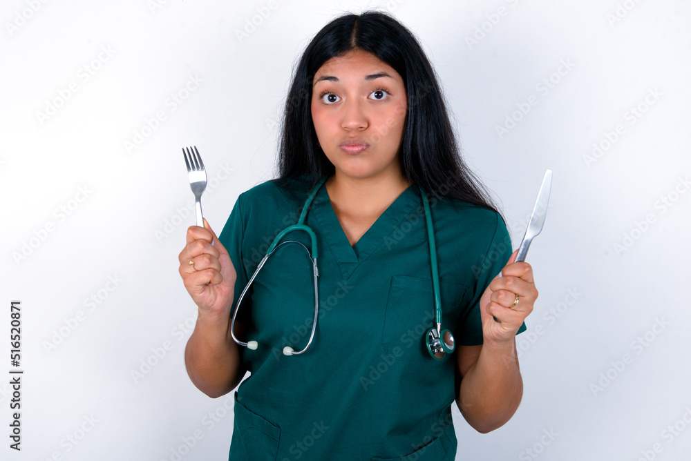 hungry Doctor hispanic woman wearing surgeon uniform over white wall holding in hand fork knife want tasty yummy pizza pie hungry Doctor hispanic woman wearing surgeon uniform over white wall holding in hand fork knife want tasty yummy pizza pie