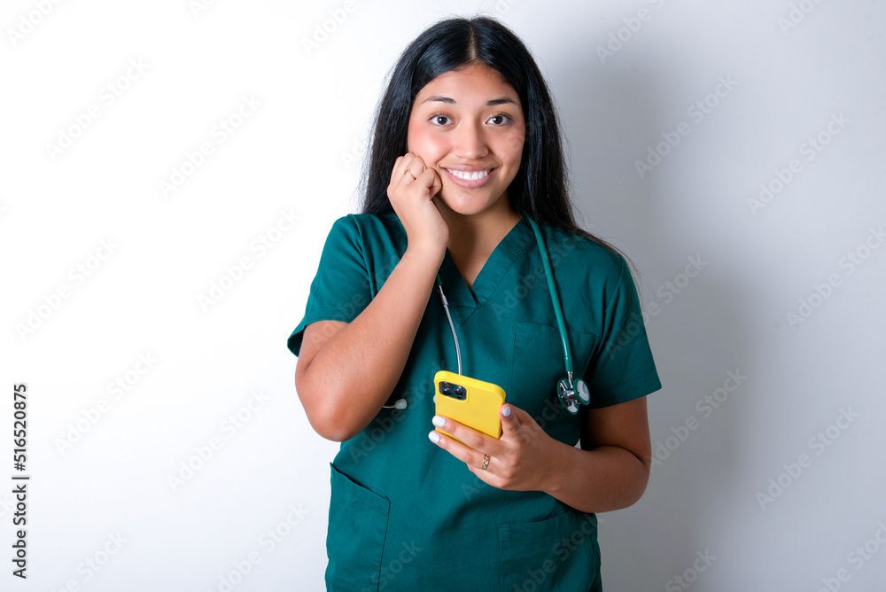Joyous Doctor hispanic woman wearing surgeon uniform poses with mobile phone device, types text message on modern smartphone, watches funny video during free time, enjoys good internet connection, Joyous Doctor hispanic woman wearing surgeon uniform poses with mobile phone device, types text message on modern smartphone, watches funny video during free time, enjoys good internet connection,
