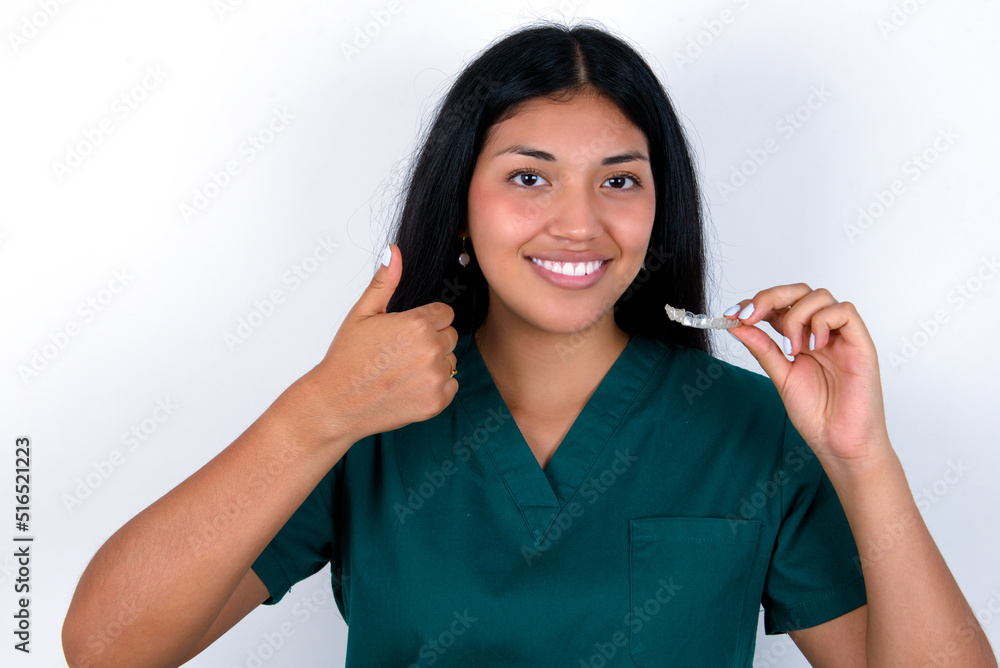 Doctor hispanic woman wearing surgeon uniform over white wall holding an invisible braces aligner and rising thumb up, recommending this new treatment. Dental healthcare concept. Doctor hispanic woman wearing surgeon uniform over white wall holding an invisible braces aligner and rising thumb up, recommending this new treatment. Dental healthcare concept.