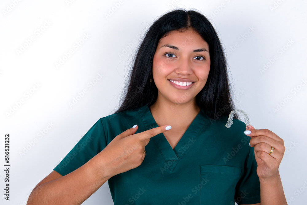 Doctor hispanic woman wearing surgeon uniform over white wall holding an invisible aligner and pointing at it. Dental healthcare and confidence concept. Doctor hispanic woman wearing surgeon uniform over white wall holding an invisible aligner and pointing at it. Dental healthcare and confidence concept.