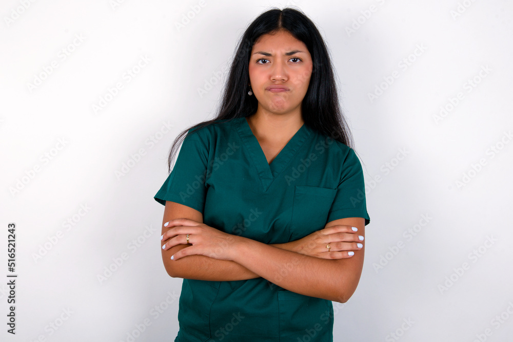 Fototapeta premium Gloomy dissatisfied Doctor hispanic woman wearing surgeon uniform over white wall looks with miserable expression at camera from under forehead, makes unhappy grimace