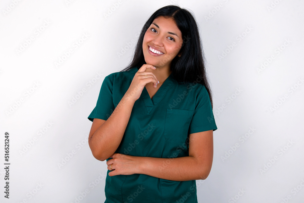 Doctor hispanic woman wearing surgeon uniform over white wall laughs happily keeps hand on chin expresses positive emotions smiles broadly has carefree expression Doctor hispanic woman wearing surgeon uniform over white wall laughs happily keeps hand on chin expresses positive emotions smiles broadly has carefree expression