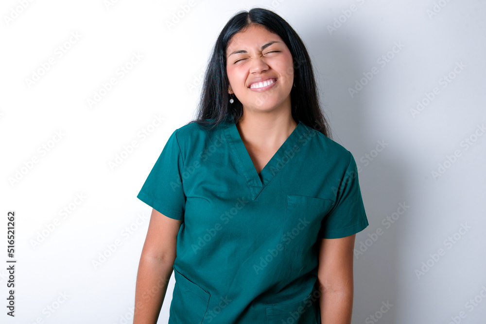 Positive Doctor hispanic woman wearing surgeon uniform over white wall with overjoyed expression closes eyes and laughs shows white perfect teeth Positive Doctor hispanic woman wearing surgeon uniform over white wall with overjoyed expression closes eyes and laughs shows white perfect teeth