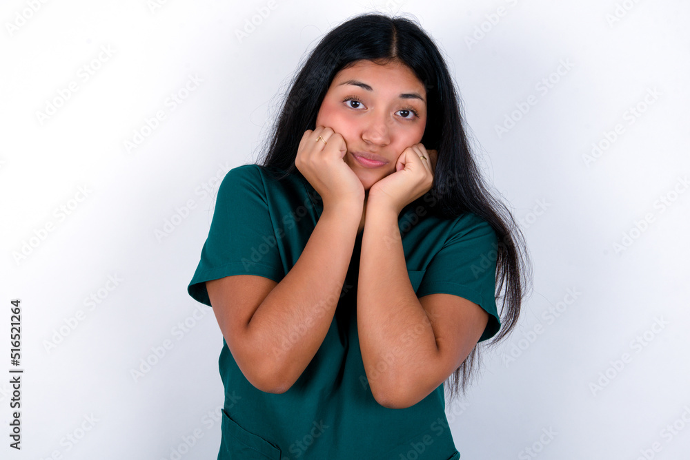 Doctor hispanic woman wearing surgeon uniform over white wall with surprised expression keeps hands under chin keeps lips folded makes funny grimace Doctor hispanic woman wearing surgeon uniform over white wall with surprised expression keeps hands under chin keeps lips folded makes funny grimace