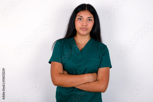 Self confident serious calm Doctor hispanic woman wearing surgeon uniform over white wall stands with arms folded. Shows professional vibe stands in assertive pose.