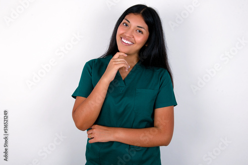 Doctor hispanic woman wearing surgeon uniform over white wall laughs happily keeps hand on chin expresses positive emotions smiles broadly has carefree expression