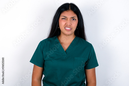 Portrait of dissatisfied Doctor hispanic woman wearing surgeon uniform over white wall smirks face, purses lips and looks with annoyance at camera, discontent hearing something unpleasant © Roquillo