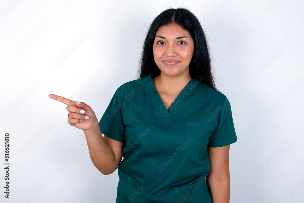Doctor hispanic woman wearing surgeon uniform over white background points to side on blank space demonstrates advertisement. People and promotion concept Doctor hispanic woman wearing surgeon uniform over white background points to side on blank space demonstrates advertisement. People and promotion concept