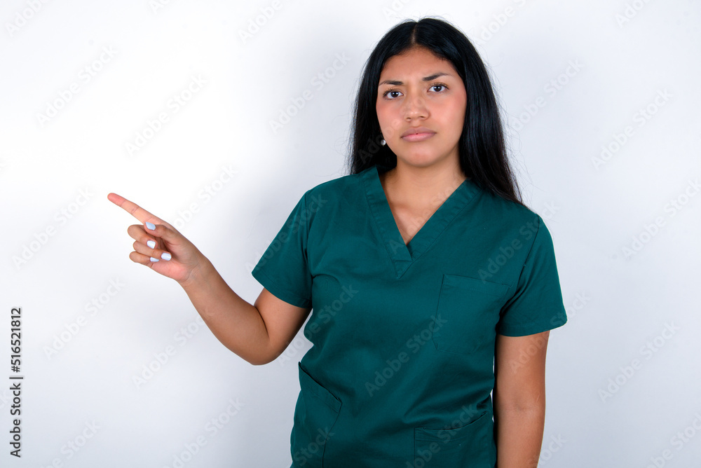 Serious Doctor hispanic woman wearing surgeon uniform over white background smirks face points away on copy space shows something unpleasant. Look at this advertisement. Big price concept Serious Doctor hispanic woman wearing surgeon uniform over white background smirks face points away on copy space shows something unpleasant. Look at this advertisement. Big price concept