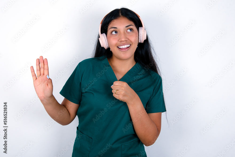 Happy Doctor hispanic woman wearing surgeon uniform over white background sings favourite song keeps hand near mouth as if microphone wears wireless headphones, listens music Happy Doctor hispanic woman wearing surgeon uniform over white background sings favourite song keeps hand near mouth as if microphone wears wireless headphones, listens music