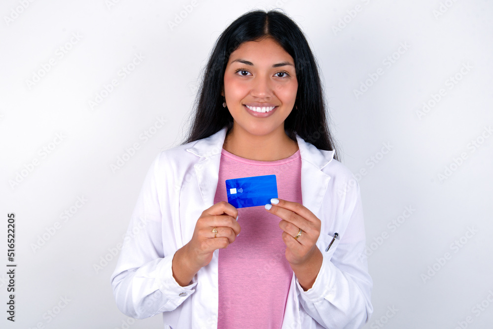 Photo of Young hispanic doctor girl wearing coat over white background positive smile hold credit card income salary Photo of Young hispanic doctor girl wearing coat over white background positive smile hold credit card income salary
