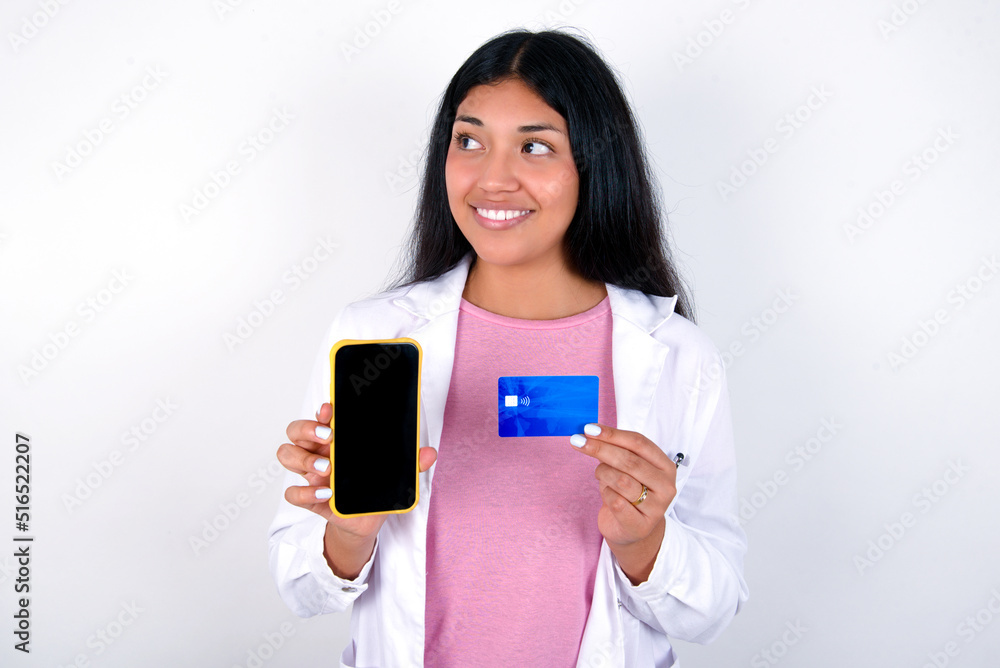 Young hispanic doctor girl wearing coat over white background holding bank card modern device looking empty space Young hispanic doctor girl wearing coat over white background holding bank card modern device looking empty space