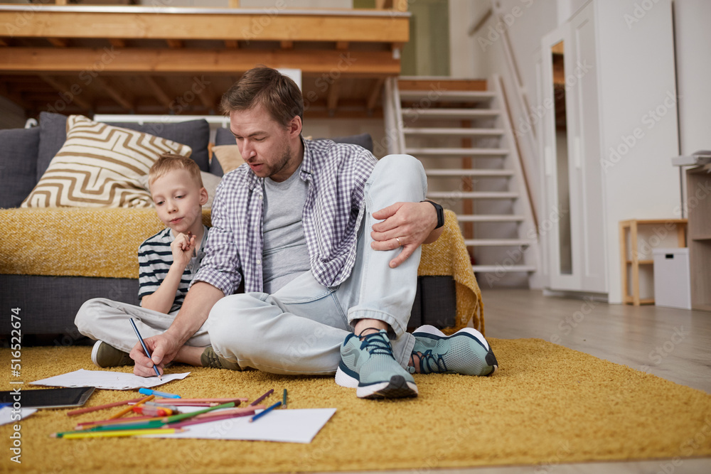Fototapeta premium Serious creative young father in casual outfit sitting on carpet and drawing with colorful pencils together with son