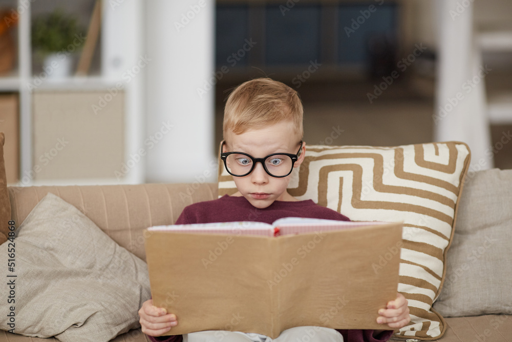 Shocked curious little boy in eyeglasses sitting on sofa and reading book at home Shocked curious little boy in eyeglasses sitting on sofa and reading book at home
