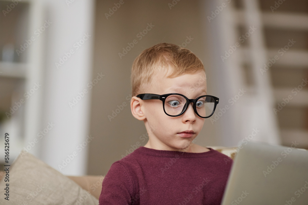 Surprised blond-haired boy in sweater and glasses sitting in living room and seeing internet advertising message Surprised blond-haired boy in sweater and glasses sitting in living room and seeing internet advertising message