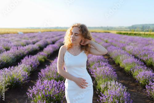 Portrait of a red-haired woman in a white dress fixing her hair and posing on a lavender field.