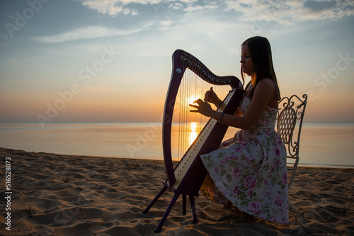 Murais de parede A girl in a flower dress plays on a Celtic harp by the sea at sunset