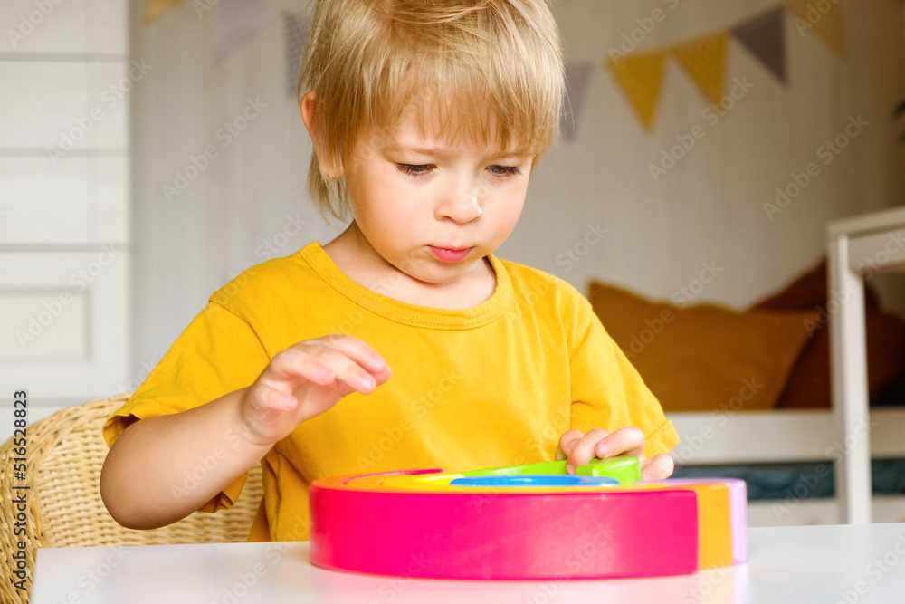 Little blond hair boy in clothes made of natural fabric plays with rainbow colored wooden toys at white table. Cozy room background in scandinavian style. Warm natural colors. Eco friendly lifestyle Little blond hair boy in clothes made of natural fabric plays with rainbow colored wooden toys at white table. Cozy room background in scandinavian style. Warm natural colors. Eco friendly lifestyle