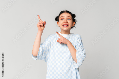 Portrait of beautiful and confident dark-haired teen girl  pointing fingers left at copy space  showing logo banner and smiling  standing in trendy t shirt over light grey background