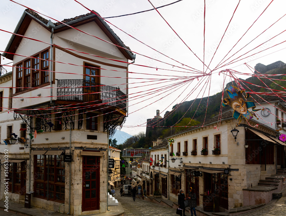 GJIROKASTER, ALBANIA - MARCH 28, 2022: Cobblestone street in historic center of city with traditional spring festive decorations and peculiar Ottoman era houses on background of hilltop castle tower