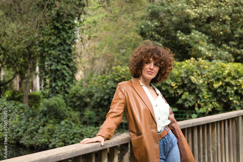 Mature and attractive woman, with curly brown hair, wearing a brown leather jacket, shirt and jeans, leaning on a wooden railing in a sexy attitude. Concept fashion, trend, beauty, sensuality, curls.