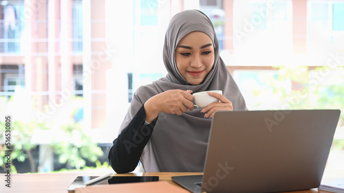Pleased muslim lady in hijab holding cup of coffee and reading business email or financial information on laptop © Prathankarnpap