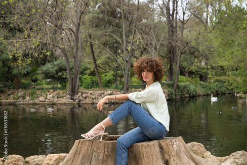 Portrait of a mature and attractive woman, with curly brown hair and light green shirt, sitting on the cut trunk of a tree by a lake. Concept beauty, fashion, trend, lake, hairstyle, curls.