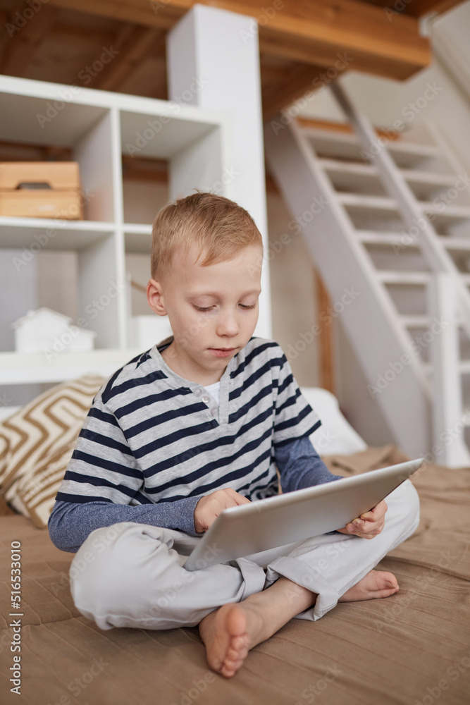 Serious blond-haired boy in casual outfit sitting with crossed legs on bed and using portable device Serious blond-haired boy in casual outfit sitting with crossed legs on bed and using portable device