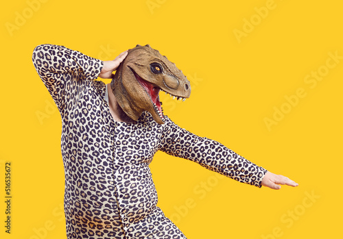 Funny eccentric Fat man dressed in leopard print pajamas dancing with dinosaur mask on his head. Cheerful crazy overweight man wearing rubber dino mask having absurd fun on orange background. Isolated photo