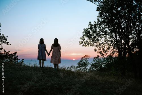 Silhouette of two teen sisters from behind holding hands and watching beautiful summer sunset over the sea 