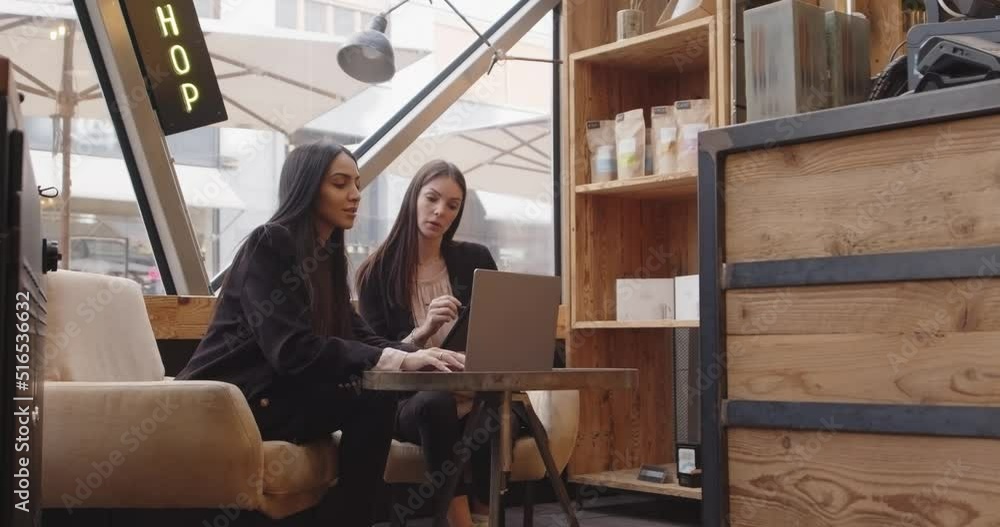 Two women sitting and working in a modern and industrial style cafe. Two multiethnic colleagues working on laptop and tablet, talking. Teamwork,corporate,diversity and social concepts Two women sitting and working in a modern and industrial style cafe. Two multiethnic colleagues working on laptop and tablet, talking. Teamwork,corporate,diversity and social concepts