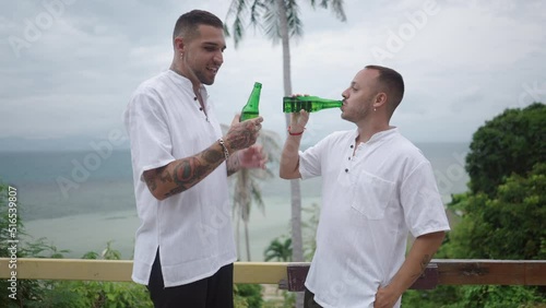 Cheerful male gay couple clinking bottles of beer while standing a tropical island terrace with a sea view  photo