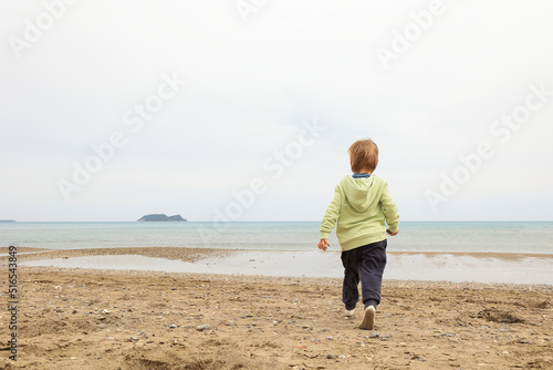 A little toddler 3years boy in yellow green cloth is walking to the sea on autumn spring beach. Cloudy day on coast. People from behind. photo