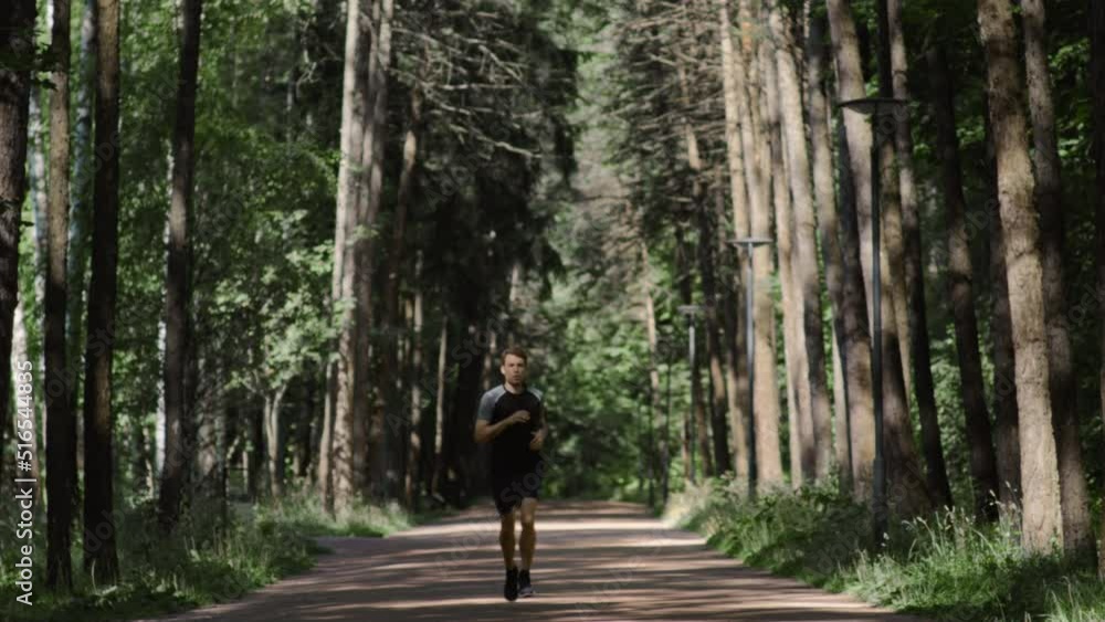 Amazing cinematic shot of handsome active fit man or athlete run, moving closer in shadowed city park or green forest. Male keeping body and mind in shape with help of smartphone health application. Amazing cinematic shot of handsome active fit man or athlete run, moving closer in shadowed city park or green forest. Male keeping body and mind in shape with help of smartphone health application.