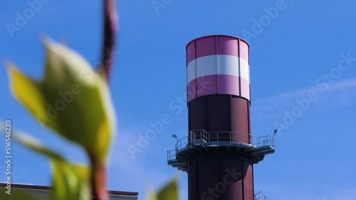 Exterior view of abandoned Soviet heavy metallurgy melting factory Liepajas Metalurgs (Liepaja, Latvia) territory, rust-covered chimney, out of focus tree in foreground, sunny day, medium shot photo