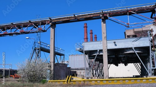 Exterior view of abandoned Soviet heavy metallurgy melting factory Liepajas Metalurgs (Liepaja, Latvia) territory, rust-covered heat pipelines, concrete poles, red brick chimneys, sunny day, wide shot photo