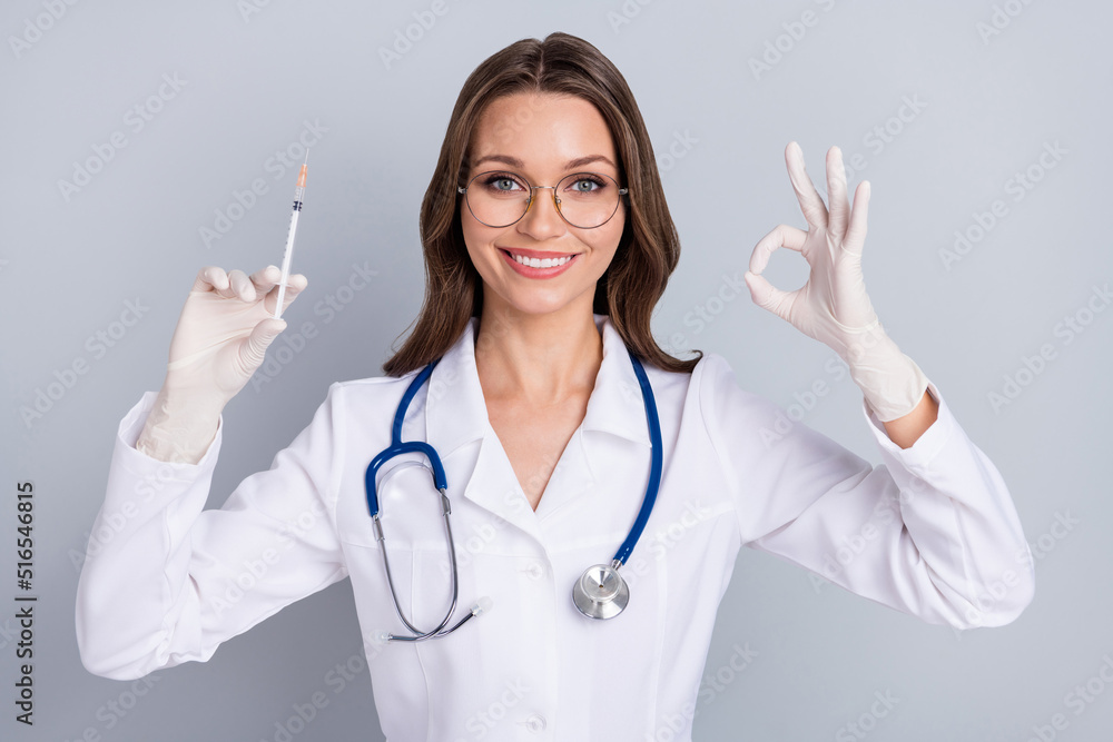 Photo of young woman pediatrician hold syringe show okey symbol good recommend isolated over grey color background Photo of young woman pediatrician hold syringe show okey symbol good recommend isolated over grey color background