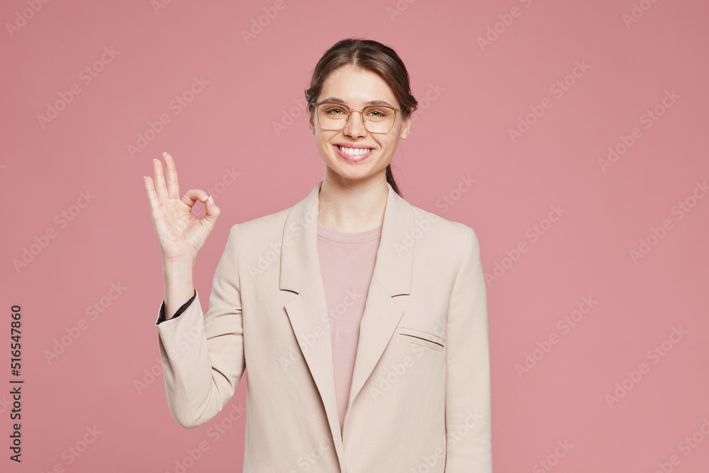 Portrait of smiling female manager in eyeglasses standing against pink background and showing ok sign while having great solution Portrait of smiling female manager in eyeglasses standing against pink background and showing ok sign while having great solution