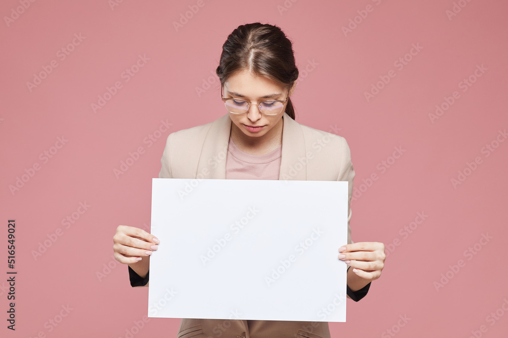 Serious smart young businesswoman in jacket standing against pink background and looking at blank poster held by her Serious smart young businesswoman in jacket standing against pink background and looking at blank poster held by her