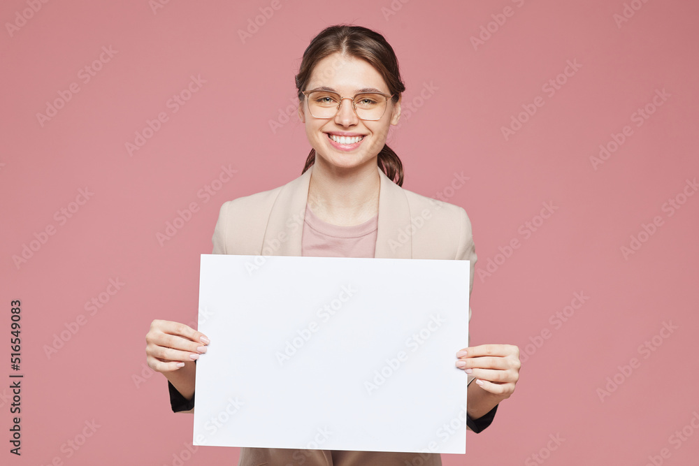 Portrait of happy beautiful intelligent young woman in eyeglasses standing against pink background and holding blank poster, advertising message concept Portrait of happy beautiful intelligent young woman in eyeglasses standing against pink background and holding blank poster, advertising message concept