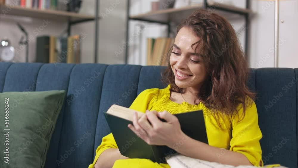 Young caucasian woman taking break from home routine and reading book while sitting on sofa at cozy home. Smiling female enjoying interesting story during weekend getaway. Young caucasian woman taking break from home routine and reading book while sitting on sofa at cozy home. Smiling female enjoying interesting story during weekend getaway.