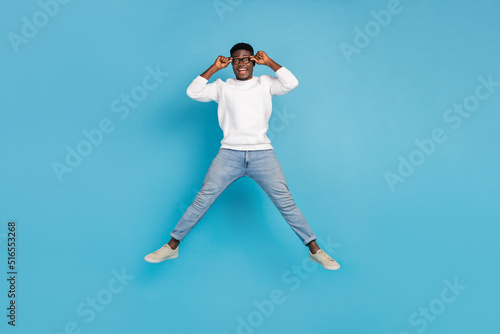 Full length portrait of astonished cheerful man jump hands touch eyeglasses isolated on blue color background