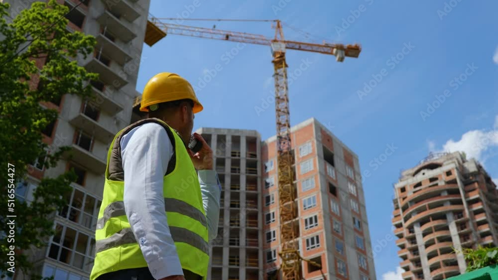 Law angle view of handsome male builder in helmet using walkie-talkie standing at construction site on street. Rear of man constructor speaking in receiver showing Up gesture Law angle view of handsome male builder in helmet using walkie-talkie standing at construction site on street. Rear of man constructor speaking in receiver showing Up gesture