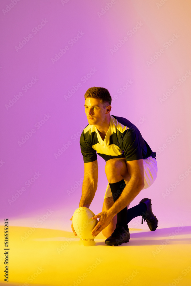 Caucasian male rugby player crouching with rugby ball over pink lighting