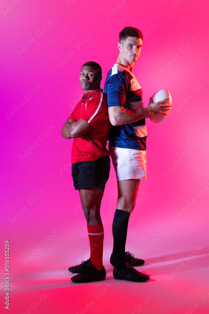 Portrait of diverse male rugby players with rugby ball over pink lighting