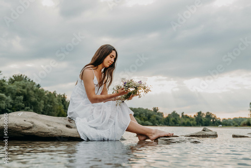 Beautiful Caucasian girl in white long dress sits on stones in middle of river and holds wreath of wildflowers in hands and looks at it. Cloudy gray sky. Woman in middle of nature. Midsummer holiday