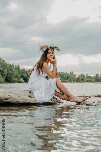 A Caucasian girl sits on a stone and has a wreath of wild flowers on her head. Man enjoys nature. Resting by the river with running water. Traditional Slavic midsummer holiday.