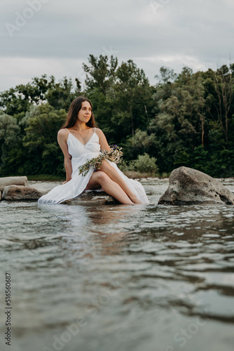A Caucasian girl with black hair sits on a stone in the water at sunset near a calm river, holding a grass wreath of wildflowers on her lap. 