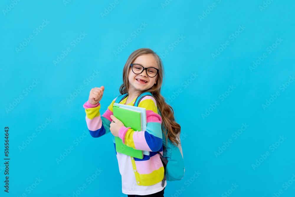 portrait of a little girl with glasses in a striped jacket with notebooks and textbooks in her hands and a backpack. The concept of education. Photo studio, blue background, place for text portrait of a little girl with glasses in a striped jacket with notebooks and textbooks in her hands and a backpack. The concept of education. Photo studio, blue background, place for text