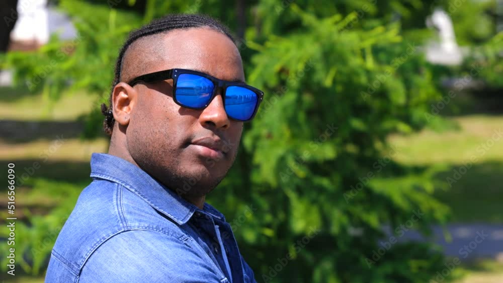 African male model in sunglasses poses against backdrop green spruce tree in city park. Stylish African-American guy in denim jacket humbly turns his head looking into camera lens of the photographer African male model in sunglasses poses against backdrop green spruce tree in city park. Stylish African-American guy in denim jacket humbly turns his head looking into camera lens of the photographer
